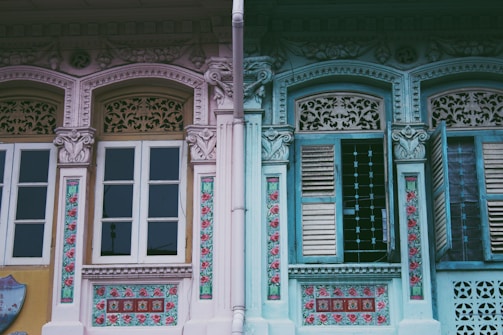 Two traditional shophouse facades with intricate details in pastel colors. The left side showcases beige and pink hues with floral designs, while the right side features turquoise and blue shades with partially open wooden shutters.
