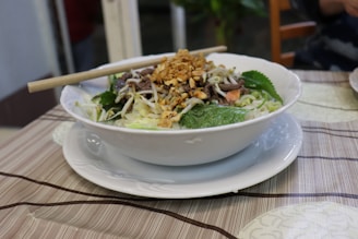 A bowl of noodles topped with sliced beef, bean sprouts, crushed peanuts, and fresh herbs sits on a white plate. The dish is served with chopsticks resting on the bowl, set on a striped tablecloth. In the background, there is a blurred view of a room with plants and furniture.