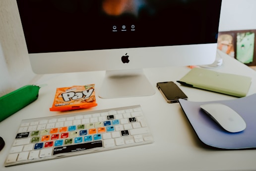A modern workspace featuring an Apple computer with a black screen, a colorful keyboard overlay, a smartphone, a mouse on a purple mouse pad, a green pencil case, and a packet of candy labeled 'Pay'. The setup is on a white desk, creating a clean and organized look.