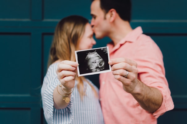 A couple is holding up an ultrasound image while standing close together. The man is kissing the woman's forehead. The focus is on the ultrasound photo, and they appear to be celebrating or sharing news about a pregnancy. They are standing in front of a dark teal wall.