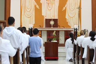 A religious ceremony takes place in a church with several people dressed in white robes kneeling in prayer. The altar is adorned with a golden monstrance, candles, and flowers. An artwork of religious figures is depicted on the wall behind.