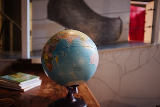 A terrestrial globe is positioned on a wooden table, with a few books stacked nearby. The globe displays detailed world geography with various countries and continents visible. In the background, there is a blurred image of a window with a red curtain or object partially visible.