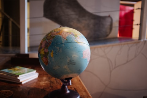 A terrestrial globe is positioned on a wooden table, with a few books stacked nearby. The globe displays detailed world geography with various countries and continents visible. In the background, there is a blurred image of a window with a red curtain or object partially visible.