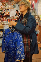 A person wearing glasses and a plaid scarf is examining a blue garment with floral patterns, possibly in a market setting. Various colorful items and decorations are visible in the background.