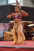 A woman dressed in a colorful traditional costume is performing a dance on stage. She wears a bejeweled headpiece, and her clothing is adorned with intricate patterns and embellishments. The stage is set with musical instruments, including large drums in the background.