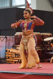 A woman dressed in a colorful traditional costume is performing a dance on stage. She wears a bejeweled headpiece, and her clothing is adorned with intricate patterns and embellishments. The stage is set with musical instruments, including large drums in the background.