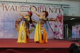 Two performers in colorful traditional costumes are dancing on a stage with a backdrop that has 'Festival dell’Oriente' written on it. They are wearing yellow skirts, red hats, and have long black hair. The stage is draped in red and the background shows decorative elements.