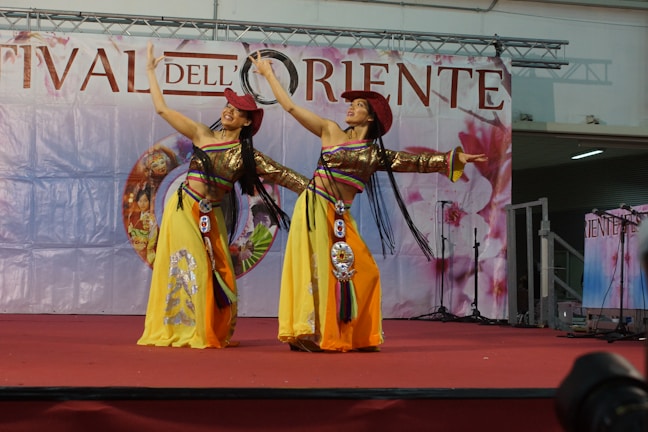 Two performers in colorful traditional costumes are dancing on a stage with a backdrop that has 'Festival dell’Oriente' written on it. They are wearing yellow skirts, red hats, and have long black hair. The stage is draped in red and the background shows decorative elements.
