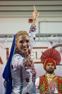 A female dancer with a bright smile is in the foreground, wearing a vibrant, colorful outfit with intricate embroidery. Her arm is extended upwards in an expressive dance pose. In the background, a male dancer is present, wearing a traditional turban and a multicolored outfit.