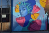 A colorful mural featuring a stylized portrait of a person with a blue face. Surrounding the face are large, vibrant flowers in shades of pink, yellow, and orange, and green leaves. The mural is painted on a large wall next to a window with wooden panels.