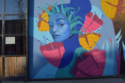 A colorful mural featuring a stylized portrait of a person with a blue face. Surrounding the face are large, vibrant flowers in shades of pink, yellow, and orange, and green leaves. The mural is painted on a large wall next to a window with wooden panels.