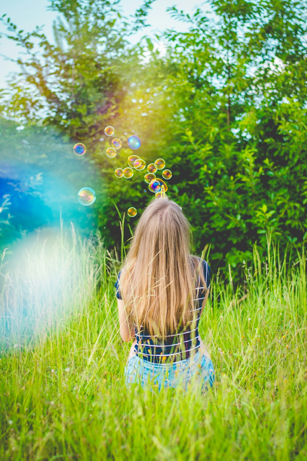 낮에 푸른 잎 나무 근처의 푸른 잔디밭에서 거품 놀이를 하면서 앉아있는 소녀