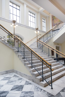 A large, elegant interior staircase made of white marble with black metal railings. Brass light fixtures hang from the ceiling, while tall windows allow natural light into the space. The marble floor below features a checkered pattern.