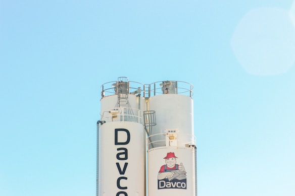 Two large industrial silos with railings and machinery at the top, set against a clear blue sky. The branding on the silo reads 'Davco' with a logo depicting a smiling character giving a thumbs-up.