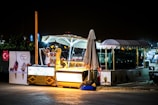 A night scene featuring an illuminated ice cream stand with an umbrella and a canopy. A person is interacting with the stand, possibly serving or setting up. The stand displays colorful advertisements with images of ice creams. Nearby, there are other structures and light sources in the background.