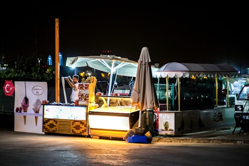A night scene featuring an illuminated ice cream stand with an umbrella and a canopy. A person is interacting with the stand, possibly serving or setting up. The stand displays colorful advertisements with images of ice creams. Nearby, there are other structures and light sources in the background.