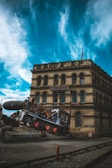 A vintage building with the sign 'Steampunk HQ' is situated under a dramatic sky filled with swirling blue and white clouds. In front of the building, an old-fashioned locomotive with a steampunk aesthetic, featuring machinery and pipes, is positioned at an angle on a rocky platform. The scene combines industrial elements with historical architecture.