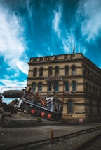 A vintage building with the sign 'Steampunk HQ' is situated under a dramatic sky filled with swirling blue and white clouds. In front of the building, an old-fashioned locomotive with a steampunk aesthetic, featuring machinery and pipes, is positioned at an angle on a rocky platform. The scene combines industrial elements with historical architecture.
