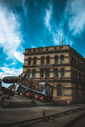 A vintage building with the sign 'Steampunk HQ' is situated under a dramatic sky filled with swirling blue and white clouds. In front of the building, an old-fashioned locomotive with a steampunk aesthetic, featuring machinery and pipes, is positioned at an angle on a rocky platform. The scene combines industrial elements with historical architecture.