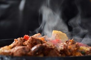 A close-up of sizzling, steaming food on a hot plate. Pieces of browned meat are mixed with colorful vegetables, including corn and red peppers. The steam rises visibly against a dark background, emphasizing the hot and fresh nature of the dish.