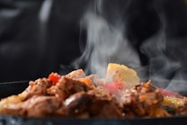 A close-up of sizzling, steaming food on a hot plate. Pieces of browned meat are mixed with colorful vegetables, including corn and red peppers. The steam rises visibly against a dark background, emphasizing the hot and fresh nature of the dish.