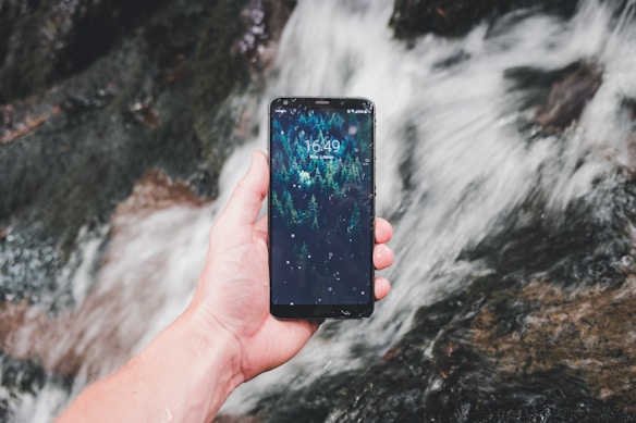A hand holding a smartphone with a forest-themed wallpaper displayed on the screen. The background consists of a flowing stream with water cascading over rocks, creating a dynamic and natural setting.