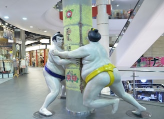 Two sumo wrestler statues seemingly engaging in a playful match, posing around a pillar in a shopping mall. The statues are painted in white with one wearing a blue mawashi and the other a yellow one. Behind them, there are various shops with merchandise displayed. The environment is brightly lit with overhead lights and there are escalators in the background.