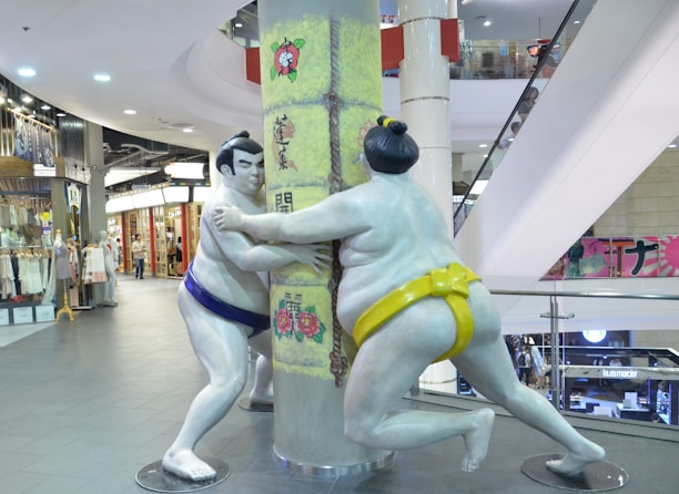 Two sumo wrestler statues seemingly engaging in a playful match, posing around a pillar in a shopping mall. The statues are painted in white with one wearing a blue mawashi and the other a yellow one. Behind them, there are various shops with merchandise displayed. The environment is brightly lit with overhead lights and there are escalators in the background.