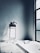 A minimalist bathroom featuring a white bathtub with a headrest and modern fixtures. The space is enhanced by a tall window on the right, allowing natural light to flood in. On the left, a small stand holds toiletries, adding a touch of practicality to the serene environment. The floor is covered with light-colored tiles.