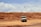 A safari vehicle is parked on a dry, grassy landscape under a partially cloudy sky. A bird is flying overhead, and the terrain is open with sparse, dried grass indicating a savanna environment.