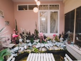 A quaint and whimsical indoor garden features pink flamingo statues and rabbit figurines amidst various plants and greenery. The background has a pink wall with a white window adorned with lace curtains. A white table is in the foreground, and koi fish swim in a small pond beneath vibrant leaf decor.