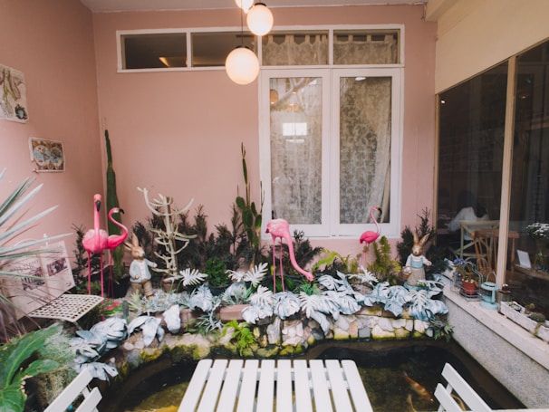 A quaint and whimsical indoor garden features pink flamingo statues and rabbit figurines amidst various plants and greenery. The background has a pink wall with a white window adorned with lace curtains. A white table is in the foreground, and koi fish swim in a small pond beneath vibrant leaf decor.