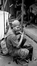 A man wearing a cap and backpack is sitting on a small stool, polishing a shoe. He is surrounded by various tools and a tin of polish. In the background, other people are sitting and engaging in conversation.