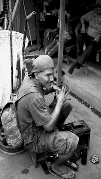 A man wearing a cap and backpack is sitting on a small stool, polishing a shoe. He is surrounded by various tools and a tin of polish. In the background, other people are sitting and engaging in conversation.