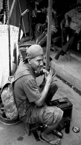 A man wearing a cap and backpack is sitting on a small stool, polishing a shoe. He is surrounded by various tools and a tin of polish. In the background, other people are sitting and engaging in conversation.