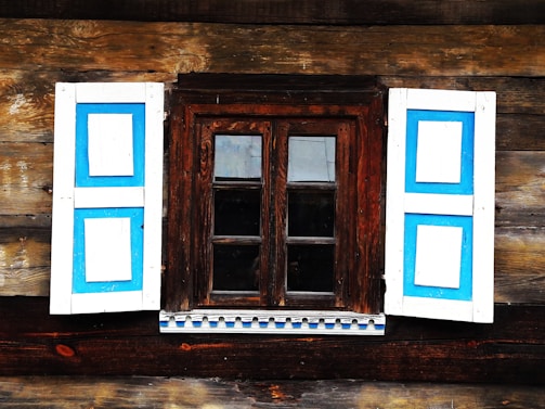 A rustic wooden wall features a small window framed by dark wood. The window has two panels, each with a distinctive blue and white shutter. The shutters are open, revealing dark reflective glass. The wood has a natural, weathered look, adding charm and character.