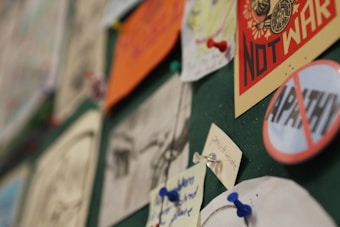 A bulletin board with various notes and posters pinned to it. The focus is on two prominent posters, one reading 'Not War' in bold red letters and another with 'Apathy' crossed out. Several smaller notes are attached with colorful pushpins.