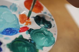 A close-up view of a painter's palette with various paint colors, including teal, orange, red, and blue. A brush is dipped into the orange paint, suggesting an ongoing art project. The palette is white and round, with wells holding the paint in place.