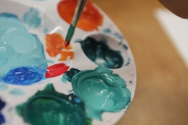 A close-up view of a painter's palette with various paint colors, including teal, orange, red, and blue. A brush is dipped into the orange paint, suggesting an ongoing art project. The palette is white and round, with wells holding the paint in place.