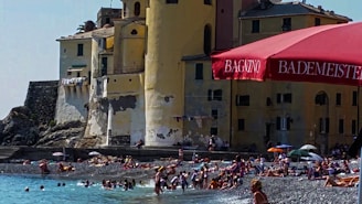 A lively beach scene with numerous people enjoying the shoreline near a historic building. The structure has aged walls and laundry hanging, giving a rustic appearance. In the foreground, a prominent red umbrella provides shade with visible text. The beach is pebbly and people are swimming and sunbathing, creating a bustling atmosphere.