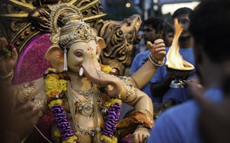 A detailed statue of Ganesha adorned with colorful garlands, surrounded by a group of people. One person holds a ceremonial flame. The statue is intricately decorated with jewelry and vibrant fabrics, set against a rich, decorative backdrop.