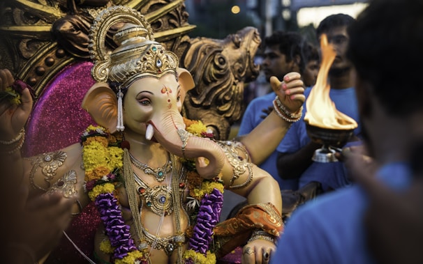 A detailed statue of Ganesha adorned with colorful garlands, surrounded by a group of people. One person holds a ceremonial flame. The statue is intricately decorated with jewelry and vibrant fabrics, set against a rich, decorative backdrop.