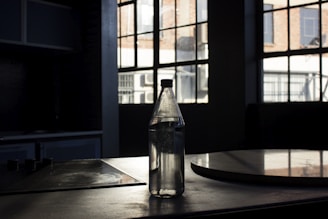 A clear water bottle is placed on a dark surface in front of large industrial-style windows that allow natural light to stream in. The setting appears to be a dimly lit room or kitchen, with visible shadows and reflections on the surface and window.