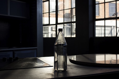 A clear water bottle is placed on a dark surface in front of large industrial-style windows that allow natural light to stream in. The setting appears to be a dimly lit room or kitchen, with visible shadows and reflections on the surface and window.