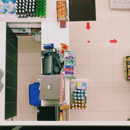 A top-down view of a supermarket checkout counter featuring various items. Eggs are in a basket at one end, and different beverages are arranged neatly on shelves, with water and soft drinks visible. A cash register and payment terminal are centrally located, surrounded by other small items like snacks and batteries. Red arrows on the floor guide customer movement.