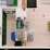 A top-down view of a supermarket checkout counter featuring various items. Eggs are in a basket at one end, and different beverages are arranged neatly on shelves, with water and soft drinks visible. A cash register and payment terminal are centrally located, surrounded by other small items like snacks and batteries. Red arrows on the floor guide customer movement.