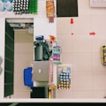 A top-down view of a supermarket checkout counter featuring various items. Eggs are in a basket at one end, and different beverages are arranged neatly on shelves, with water and soft drinks visible. A cash register and payment terminal are centrally located, surrounded by other small items like snacks and batteries. Red arrows on the floor guide customer movement.