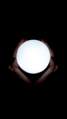 A pair of hands gently cradles a bright, glowing orb against a dark background. The light from the orb illuminates the hands, highlighting their contours and details.