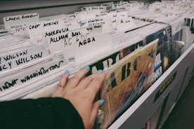 A hand with painted nails browsing through a collection of vinyl records in a store. Various artist names are written on white dividers, including 'Laurie Anderson', 'Ambrosia', 'Allman Bros.' and 'ABBA'. The records are stacked vertically in a long row, and there is a sense of nostalgia and exploration in the setting.