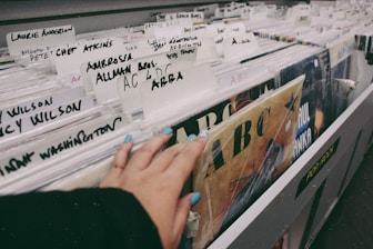 A hand with painted nails browsing through a collection of vinyl records in a store. Various artist names are written on white dividers, including 'Laurie Anderson', 'Ambrosia', 'Allman Bros.' and 'ABBA'. The records are stacked vertically in a long row, and there is a sense of nostalgia and exploration in the setting.